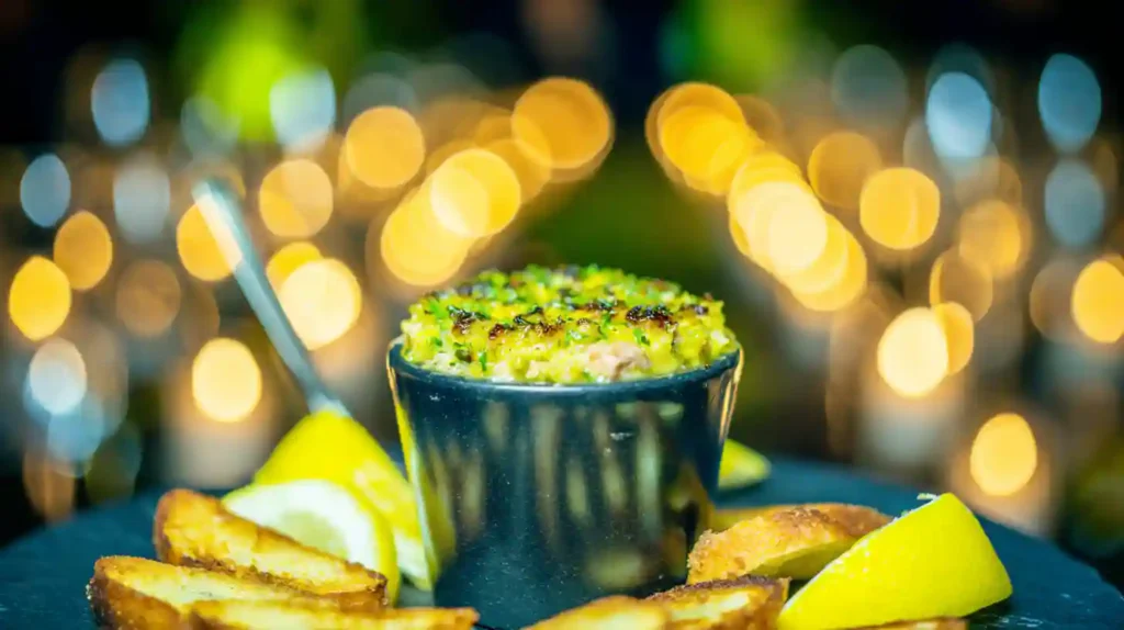 "Crab brûlée in a white ramekin with caramelized cheese topping and microgreens, served on a rustic wooden table."