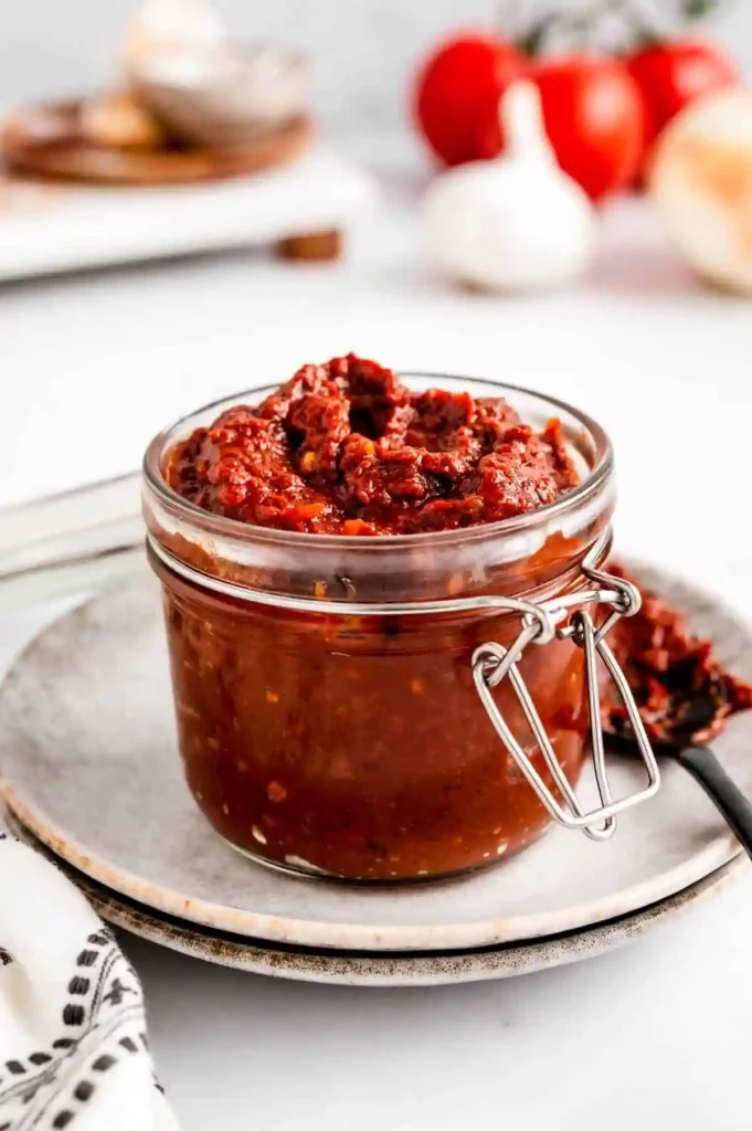 Glass jar of thick homemade Tex Mex Paste on a ceramic plate, surrounded by tomatoes, garlic, and a spoonful of paste
