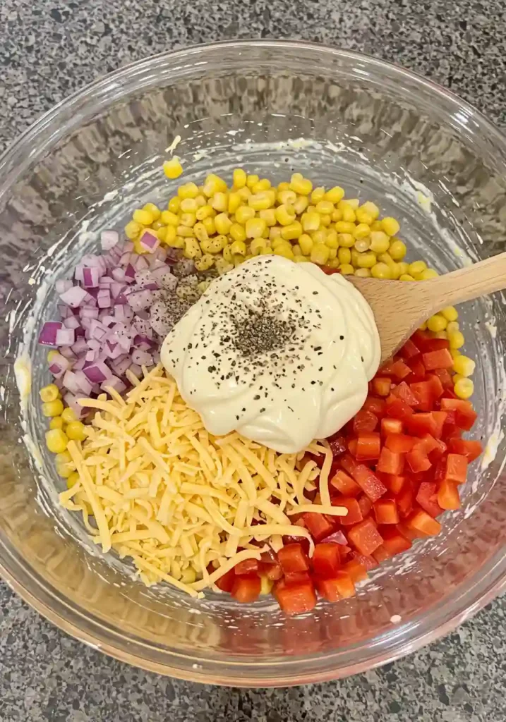 Corn, cheese, diced pepper, and mayo being mixed together in a large bowl
