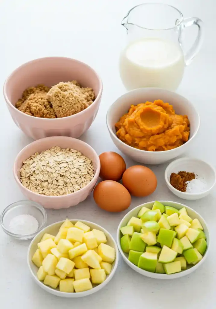 Ingredients for pumpkin apple baked oatmeal including oats, pumpkin puree, apples, eggs, and spices on white background