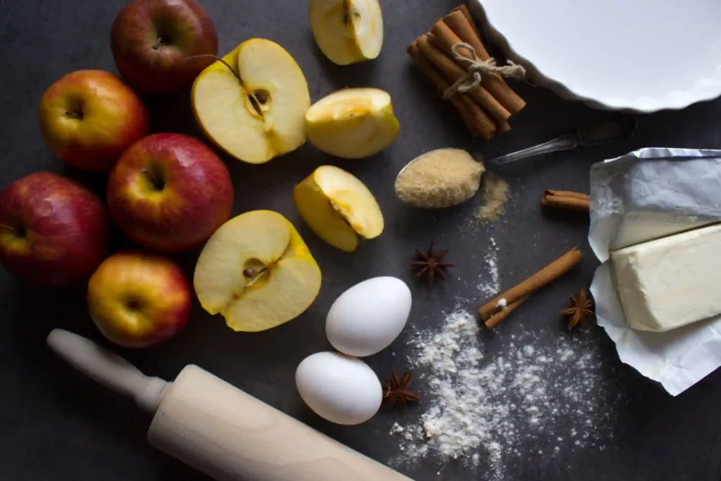 Ingredients for making an Apple Pie Recipe  laid out on a dark surface. The ingredients include fresh green apples, flour, butter, eggs, brown sugar, and cinnamon sticks, with a rolling pin and a pie dish nearby."