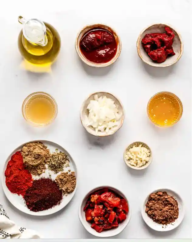 Overhead view of ingredients for homemade Tex Mex paste, including olive oil, tomato paste, chipotle in adobo, chopped onion, garlic, roasted peppers, broth, honey, and a mix of chili powder, paprika, cumin and spices on a white background
